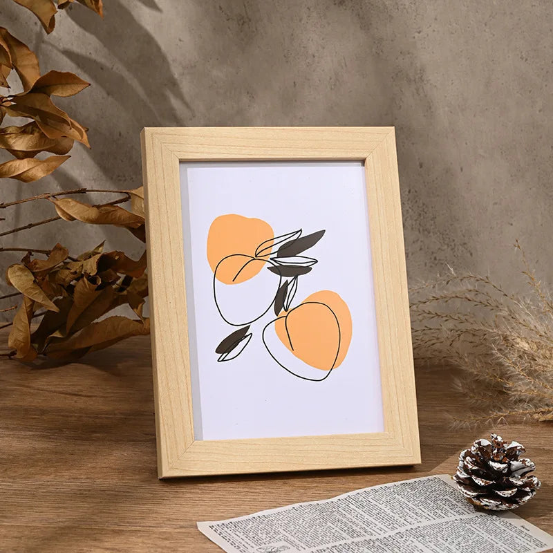 A light wooden photo frame with an orange abstract design is displayed on a wooden surface next to dried foliage.