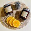 Two amber glass candle jars with metal lids and a jar of dried lavender next to sliced oranges on a woven mat.