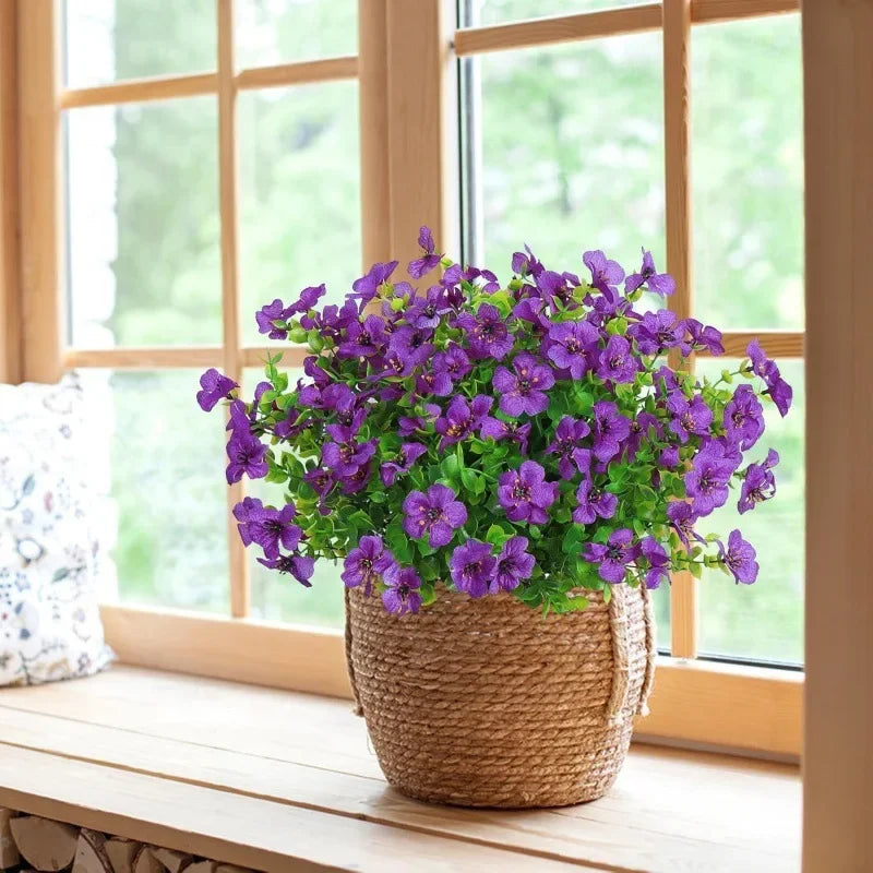 A potted arrangement of vibrant purple artificial flowers sits on a wooden windowsill next to a window.