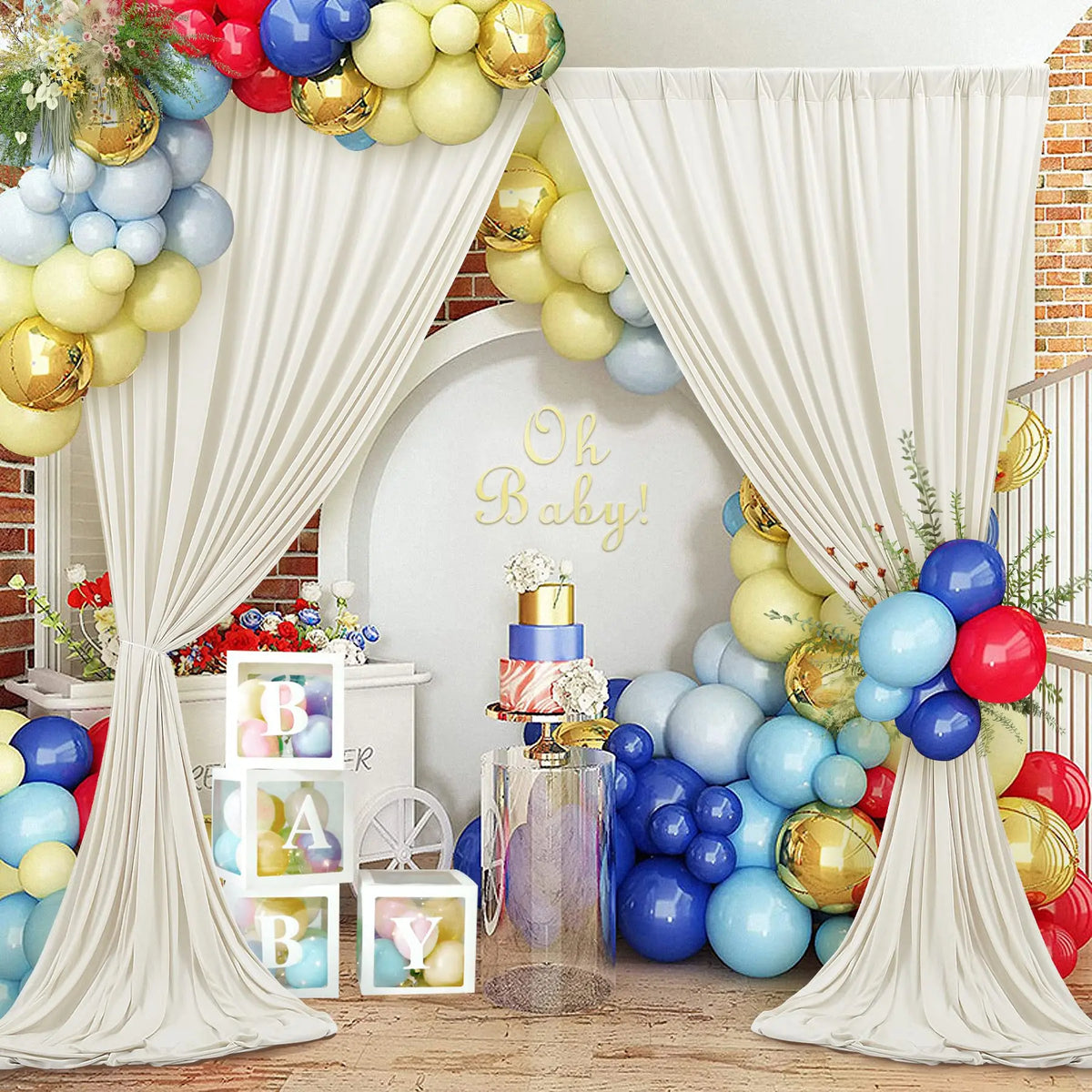 A white draped backdrop with gold balloons and blue/red balloons for a baby celebration