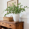 A large white ceramic vase filled with artificial eucalyptus greenery sits on a wooden dresser.