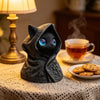 A dark gray, hooded cat statue with glowing blue eyes sits on a lace tablecloth next to a lamp.