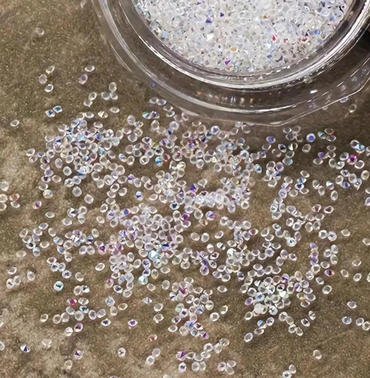 Iridescent nail rhinestones scattered on a surface next to a clear container.