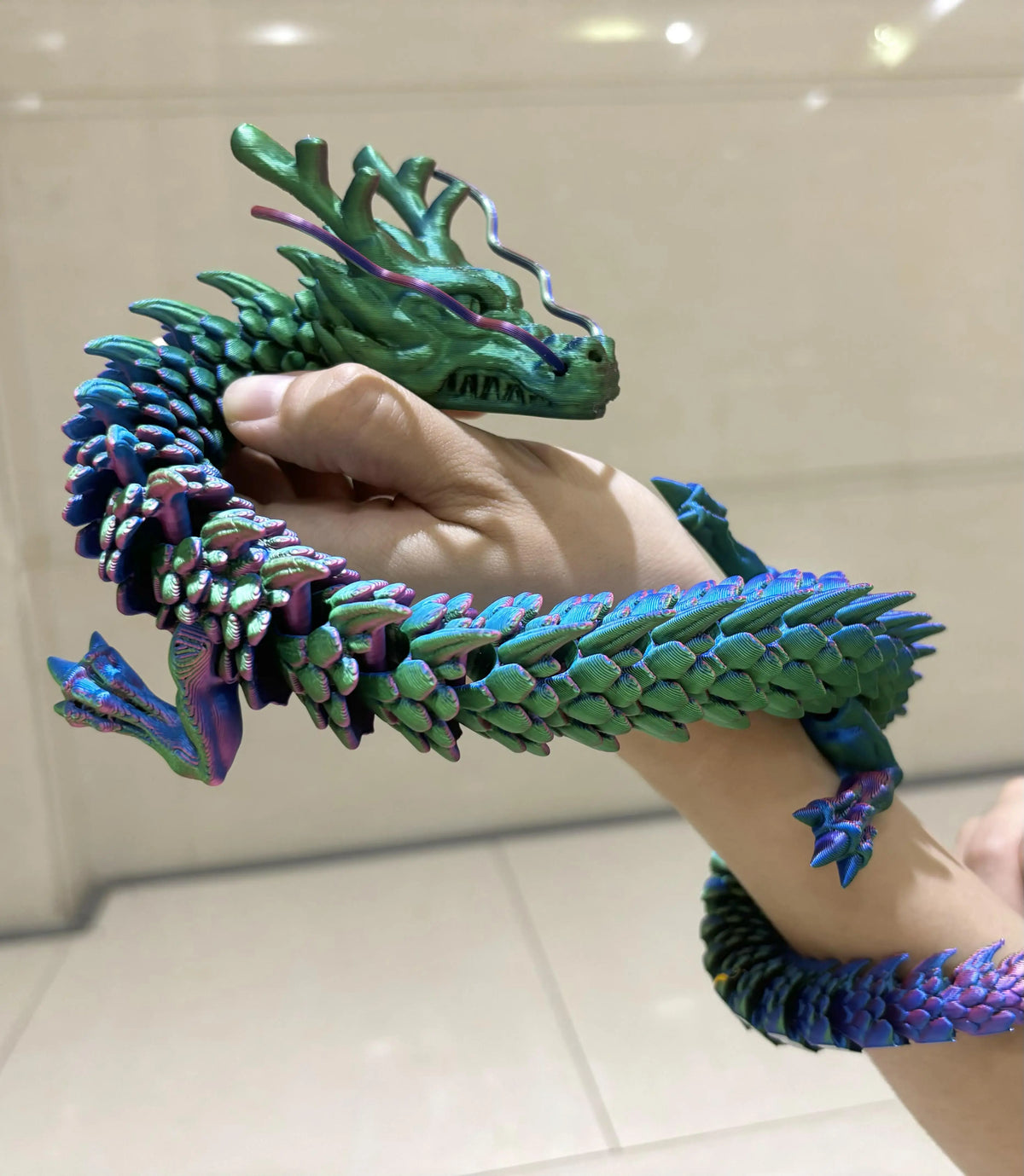 A hand holds a green and purple articulated 3D printed dragon figurine with textured scales.