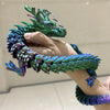 A hand holds a green and purple articulated 3D printed dragon figurine with textured scales.