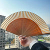 A hand holds an elegant bamboo folding fan with a classic design against a city skyline backdrop.