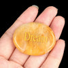 A hand holds a smooth, oval, translucent yellow-orange crystal with the word Dream engraved on it.