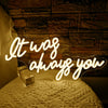 A glowing neon sign spelling out It was always you is displayed on a textured surface.