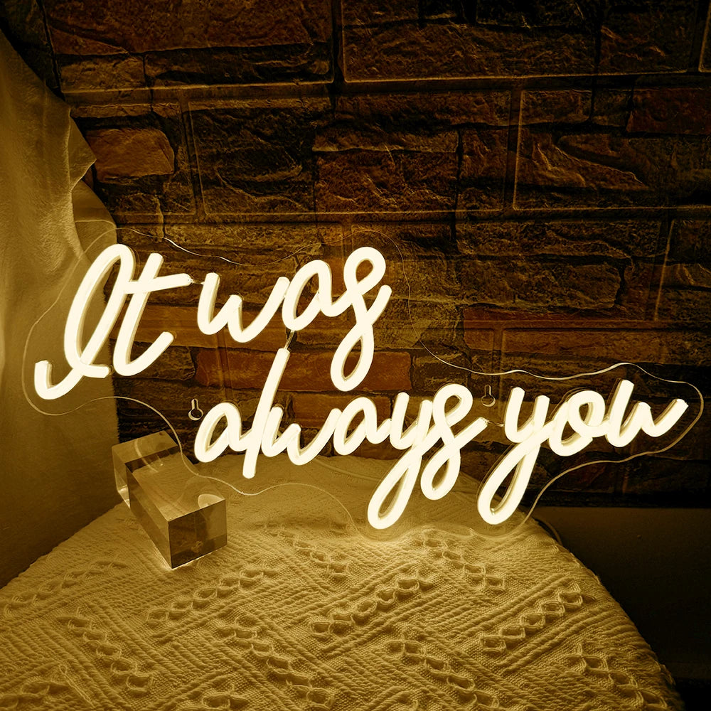 A glowing neon sign spelling out It was always you is displayed on a textured surface.