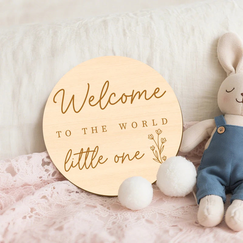 A wooden welcome sign with the text Welcome to the world little one resting on a pink lace fabric next to a white bunny figur