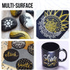 A collection of multi-surface art featuring black and gold paint pens on various surfaces like stones, paper, and a mug.
