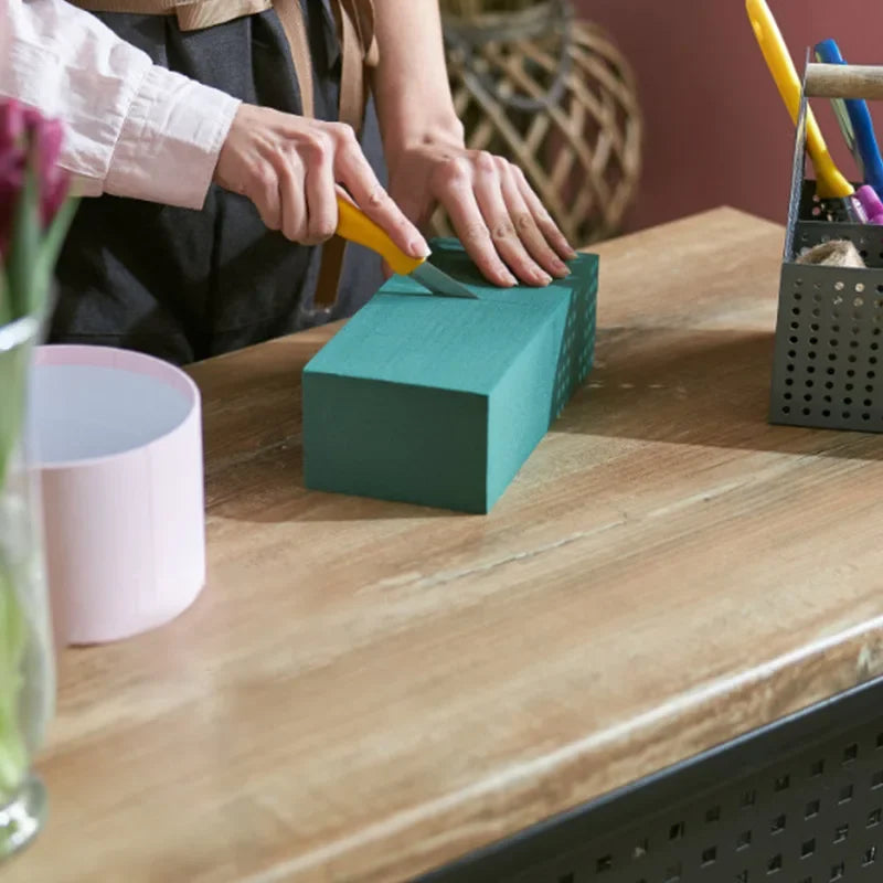 A person is cutting a teal foam block with a yellow knife on a wooden table.