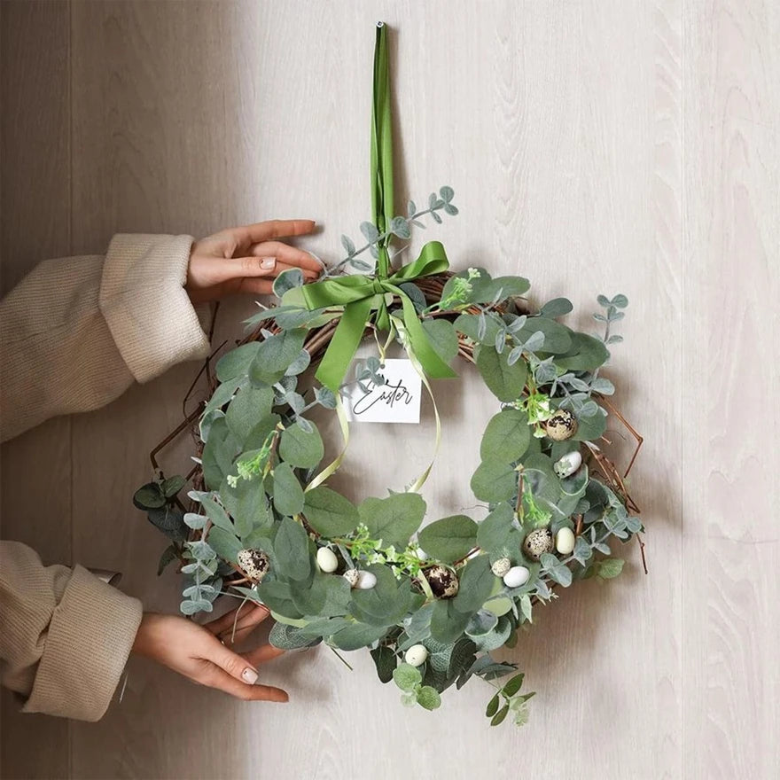 Hands hold a wreath made of artificial eucalyptus stems with small white berries and green leaves.