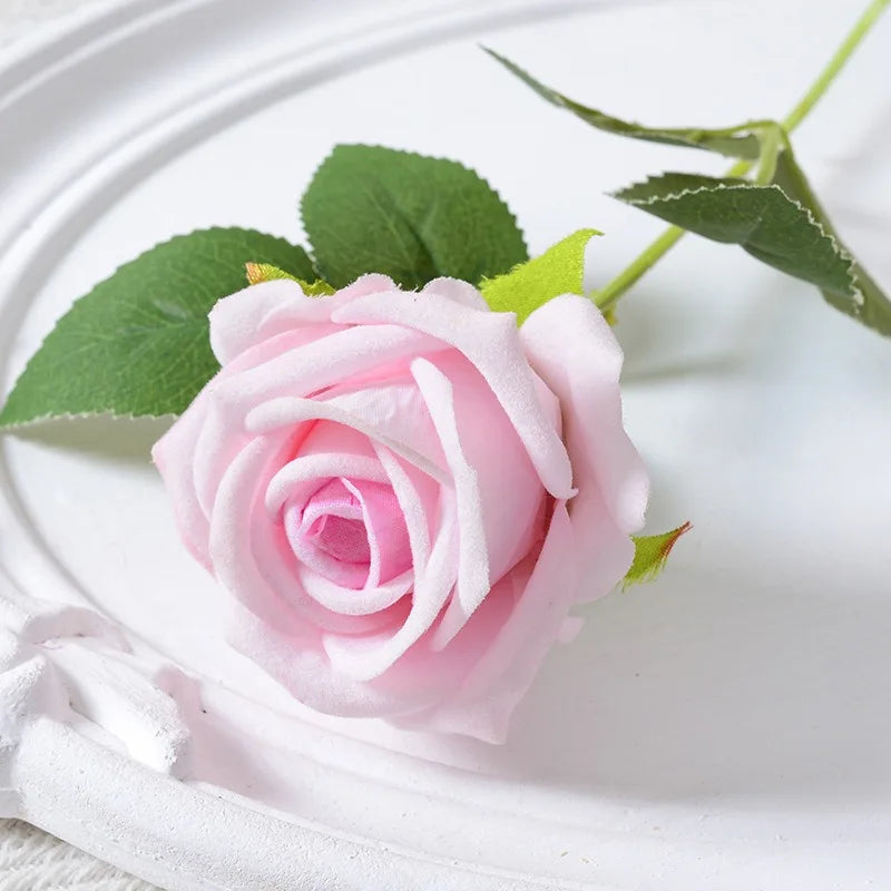 A soft pink velvet artificial rose with green leaves is displayed on a white plate.