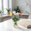 A glass vase filled with handcrafted mini glass flowers in shades of yellow, purple, and orange on a white table.