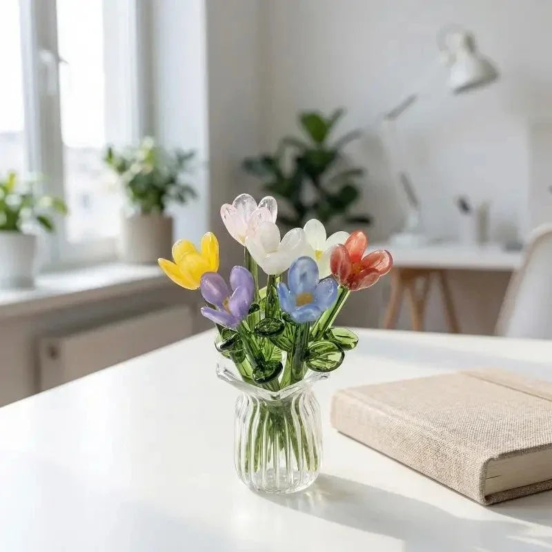 A glass vase filled with handcrafted mini glass flowers in shades of yellow, purple, and orange on a white table.