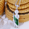 A silver pendant necklace featuring a figure and a green gemstone, displayed on a woven background.
