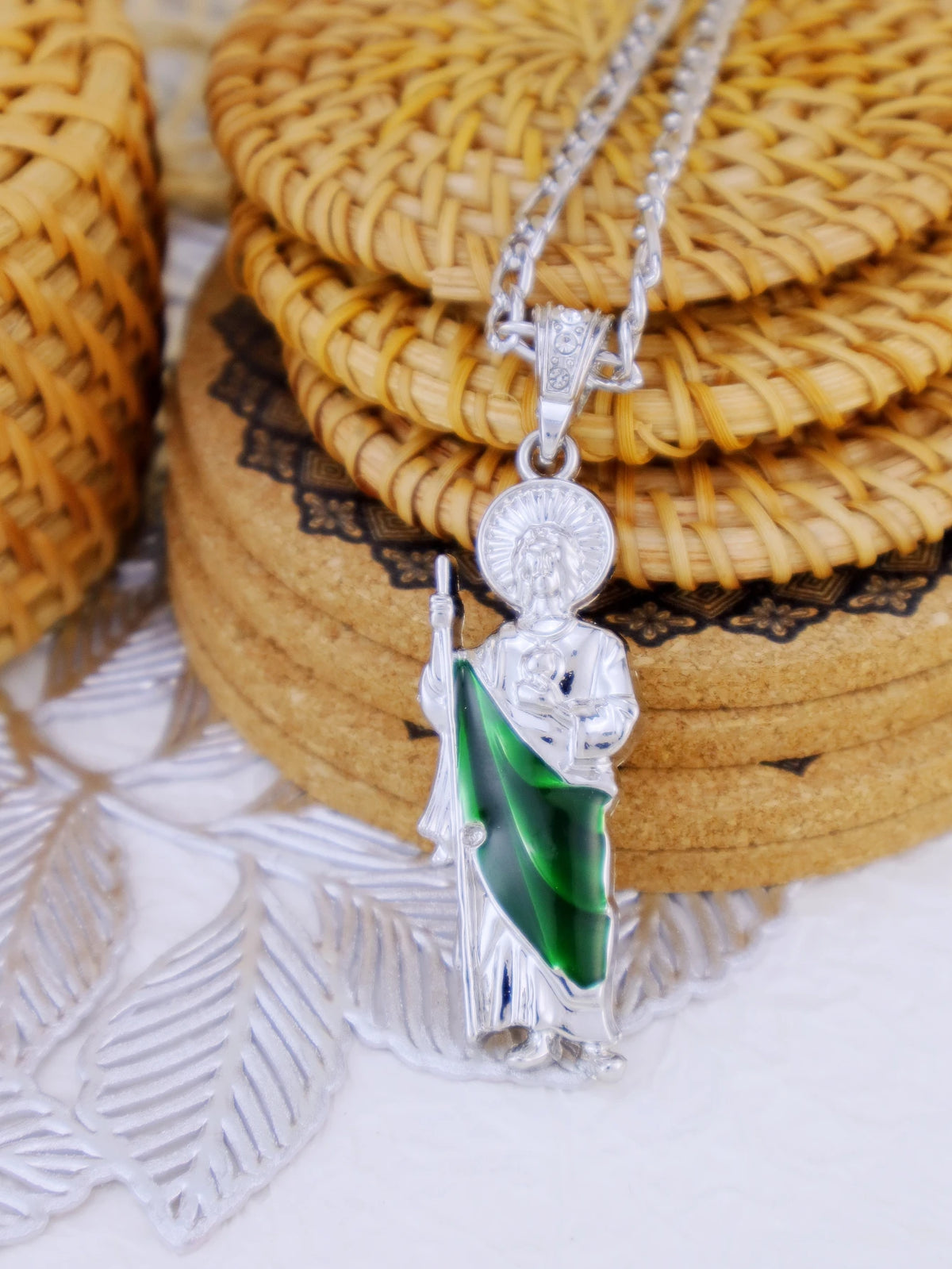 A silver pendant necklace featuring a figure and a green gemstone, displayed on a woven background.