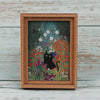 A framed painting featuring a black cat amidst colorful wildflowers and foliage.