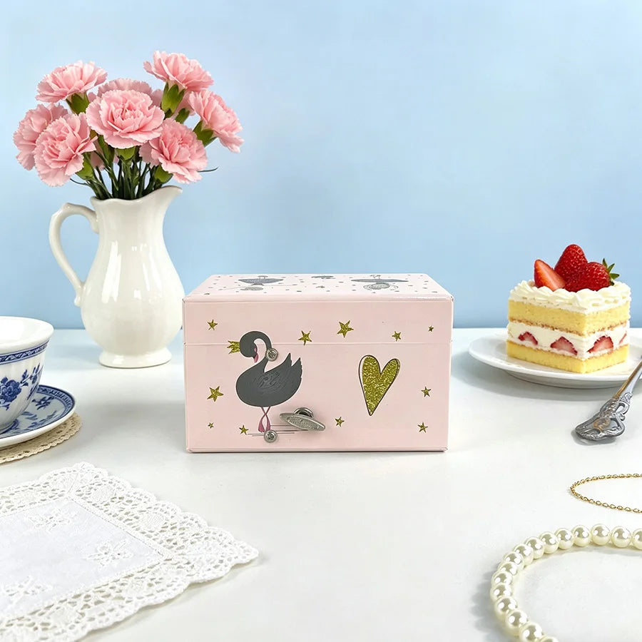 A pink ballet music box with a lid open, featuring a silhouette of a swan and a heart design, placed on a white table.