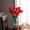 A bouquet of red roses in a glass vase with green leaves sits on a wooden surface.