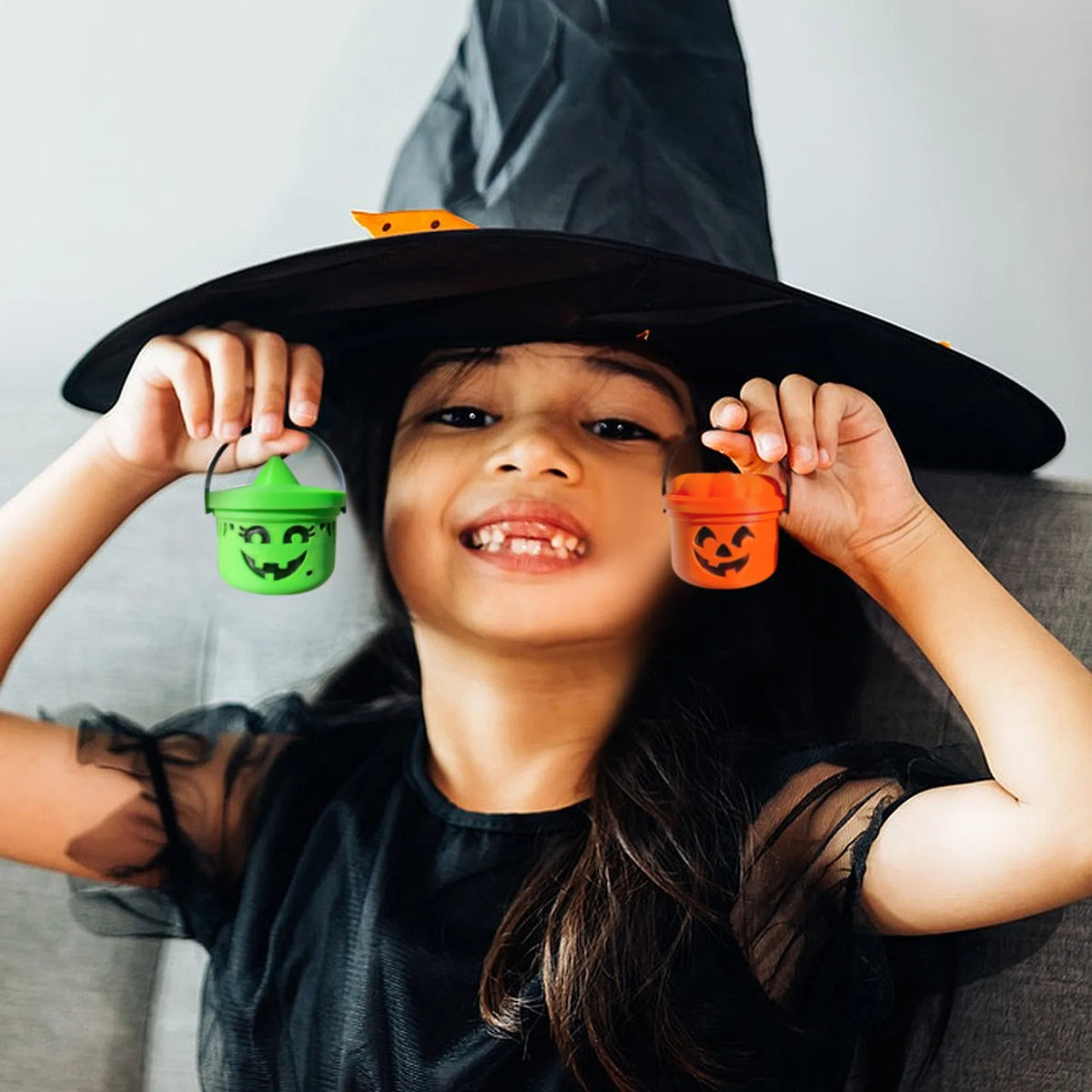 A smiling girl holds up two miniature pumpkin Halloween buckets for treats and decor.