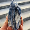 A hand holds a deep blue natural kyanite crystal stone with white inclusions against a gray striped background.