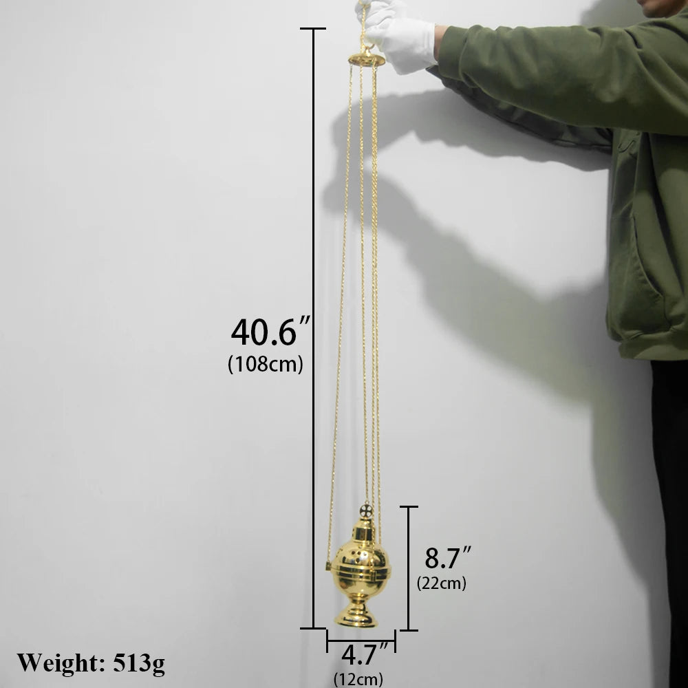 A gold-plated orthodox censer hanging by chains against a white wall, showing its dimensions.