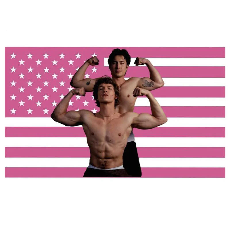Un homme musclé soulève un autre homme dans une pose devant un fond de drapeau américain rayé rose et blanc.