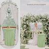 A light green stretch tablecloth draped on a round table in an outdoor wedding or event setting surrounded by white floral ar