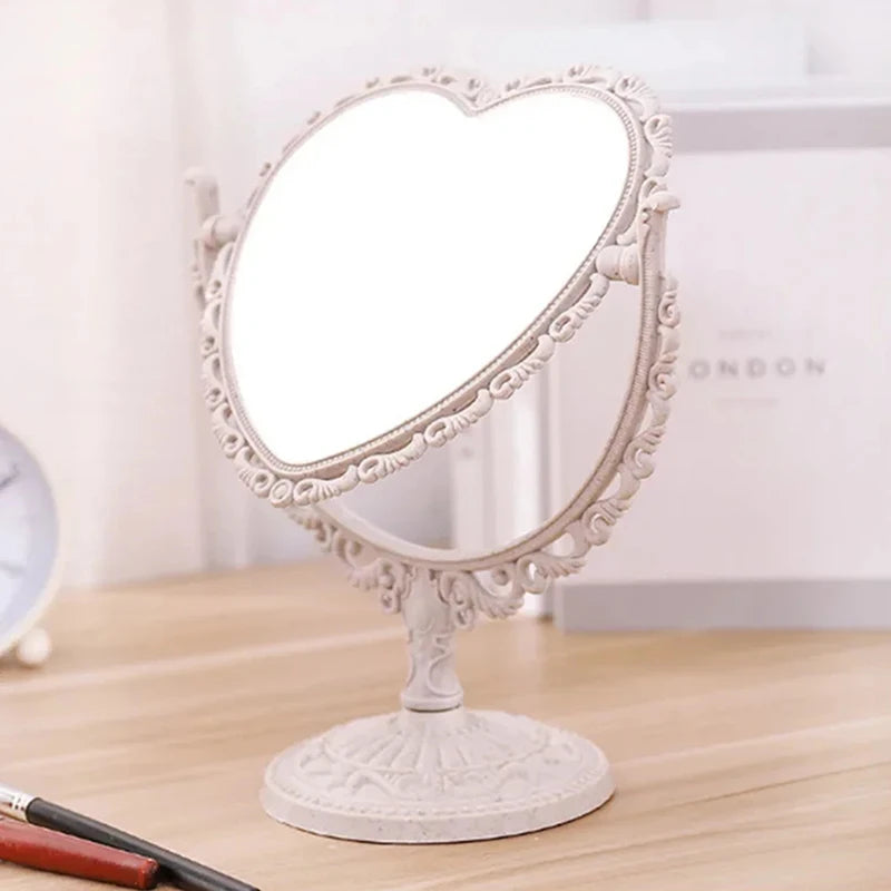 Espejo de maquillaje con forma de corazón blanco y marco decorativo sobre una superficie de madera.