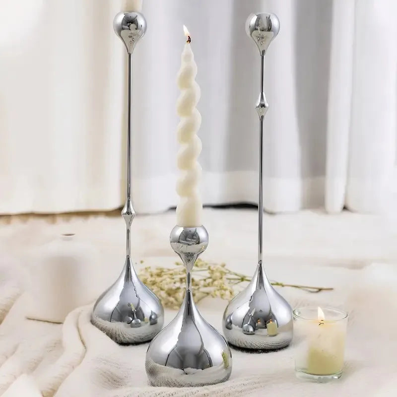 A set of three silver waterdrop design candle holders with white candles displayed on a white cloth.