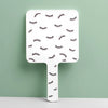 A white handheld vanity mirror with an ergonomic handle featuring a pattern of stylized eyelashes against a light green backg
