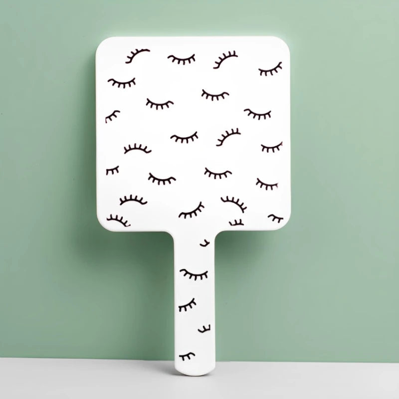 A white handheld vanity mirror with an ergonomic handle featuring a pattern of stylized eyelashes against a light green backg