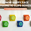 A series of colorful magnetic clips in various colors are displayed on a white refrigerator surface.