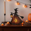 A dark metal witch ghost lantern with glowing amber light sits on a wooden surface next to candles and seasonal decorations.