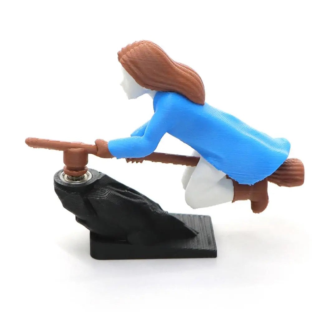 A figurine of a witch with brown hair and blue clothing is shown flying on a dashboard drift decor.