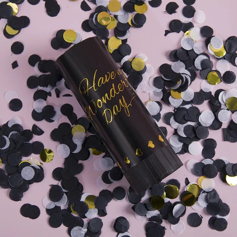 A black tube of Hand Twist Confetti Cannons lying on a pink surface surrounded by scattered black and gold confetti.