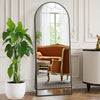 A full length body mirror with a black aluminum frame reflecting an orange velvet sofa and greenery.