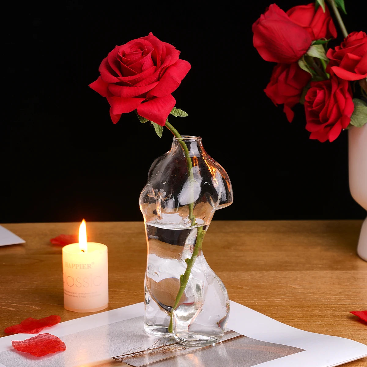 Artistic glass human body vase holding a red rose and lit candle on a wooden surface.