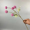 Una mano sostiene un tallo con flores artificiales de crisantemo rosa y diente de león contra un fondo gris claro.