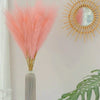 A tall vase holds a bouquet of pink pampas grass with a gold sunburst mirror decoration and green tropical leaves on a white 