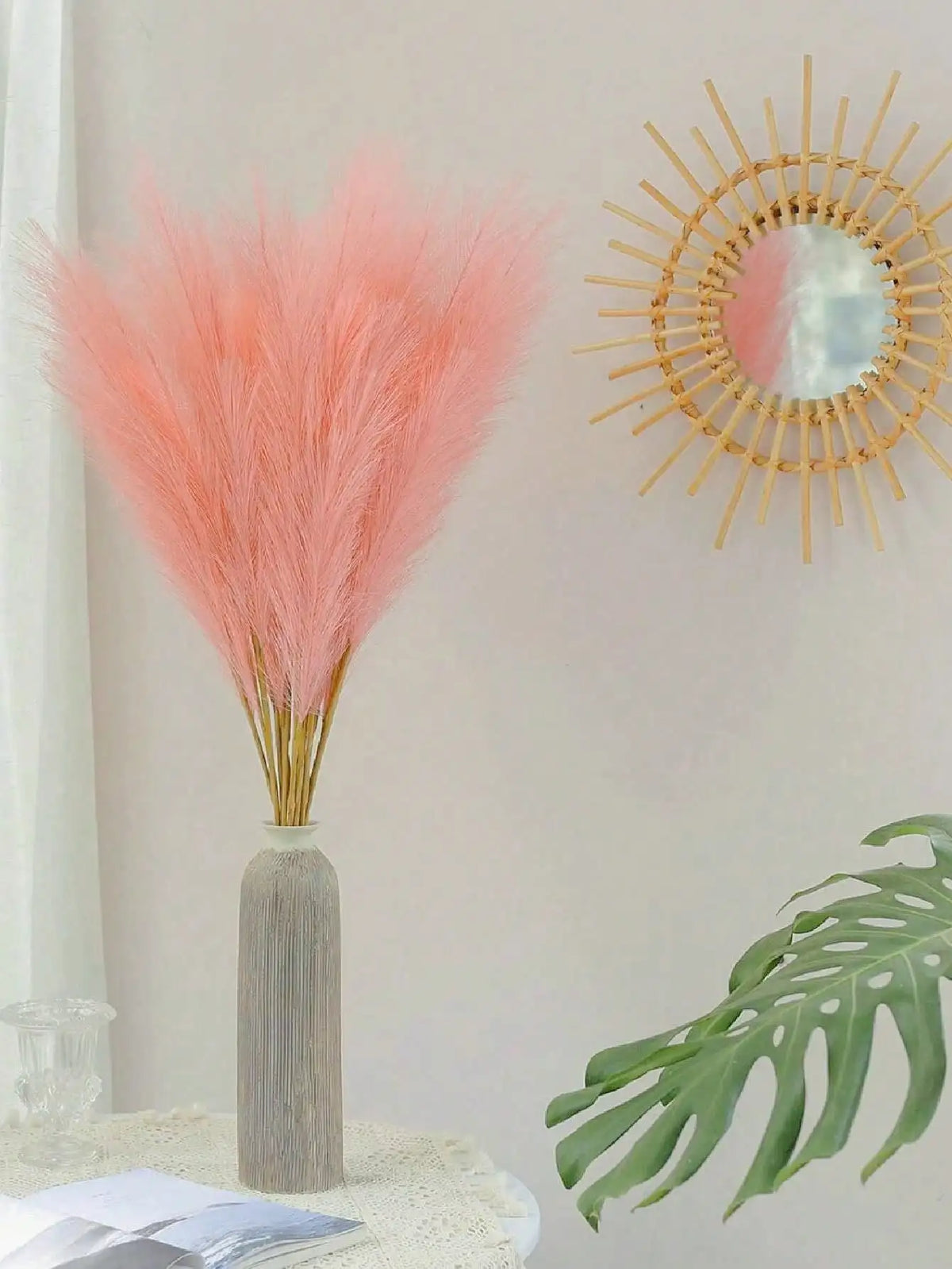 A tall vase holds a bouquet of pink pampas grass with a gold sunburst mirror decoration and green tropical leaves on a white 