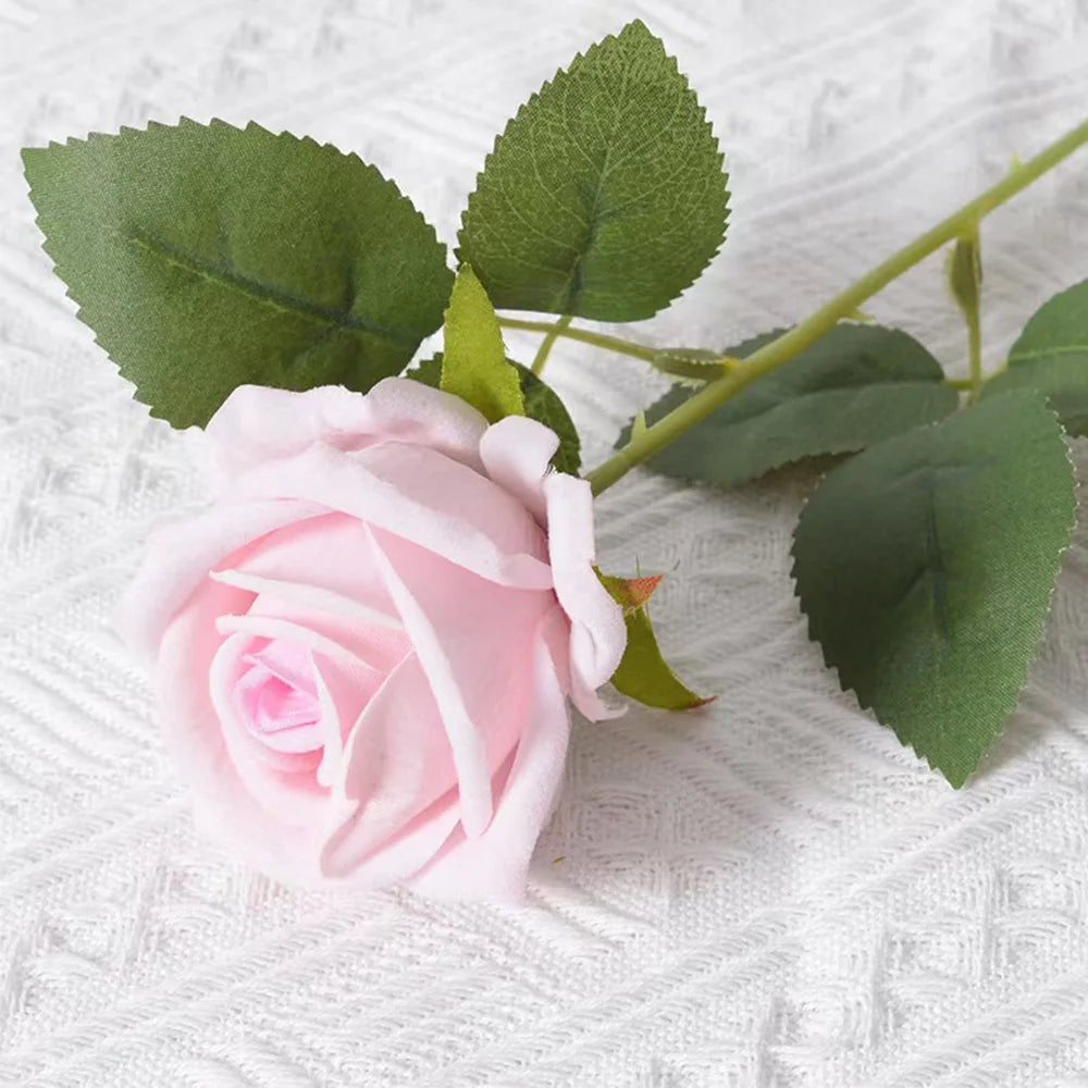 A soft pink artificial rose with green leaves on a white textured background.