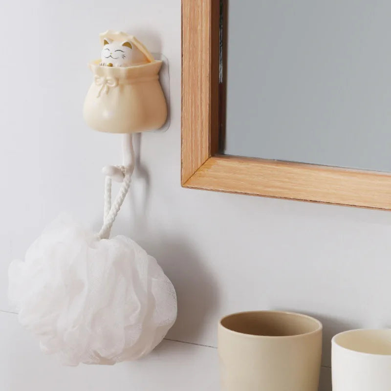 A light beige cartoon cat wall hook with a white fabric pom-pom hanging next to a wooden framed mirror.