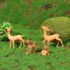 A set of small, fawn-colored deer figurines placed on green grass with a blurred green hedge background.