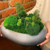 A hand holds a potted arrangement of vibrant green artificial moss plants against a brick wall.