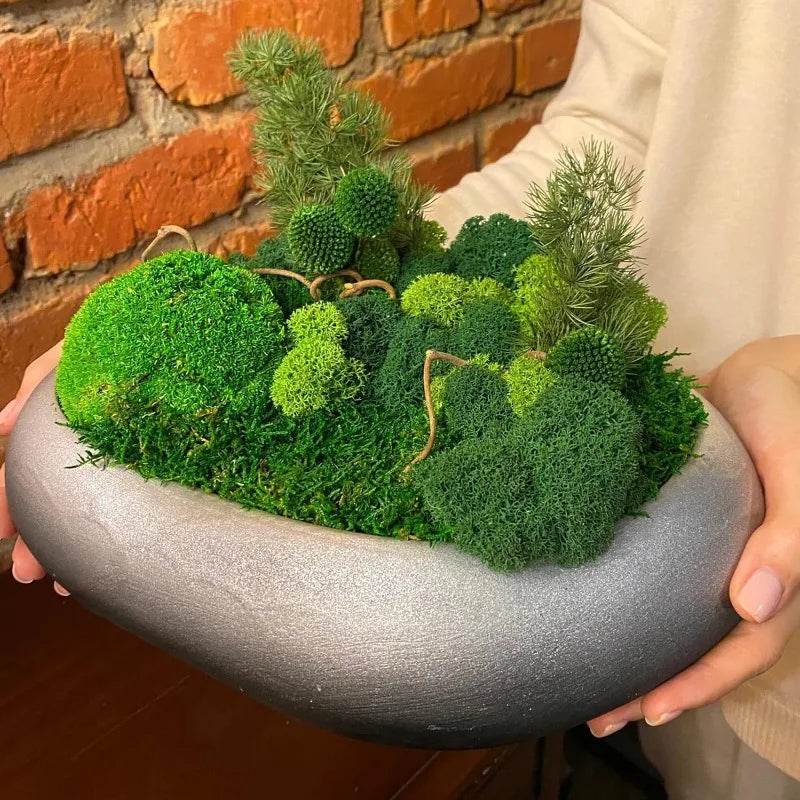 A hand holds a potted arrangement of vibrant green artificial moss plants against a brick wall.