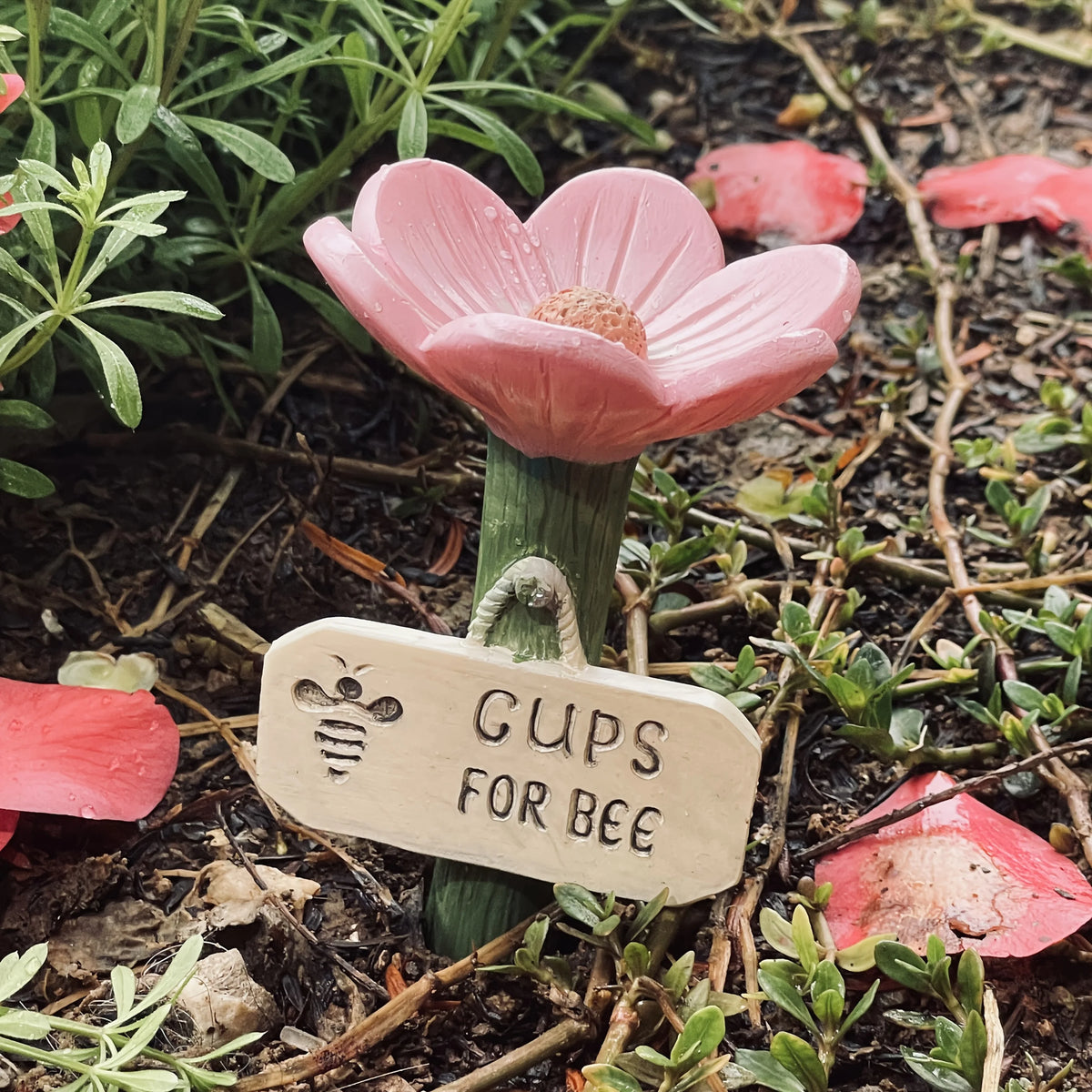 A wooden tag labeled CUPS FOR BEE is placed in the dirt next to a pink flower.