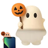 A white ghost phone decoration charm with an orange Jack-o'-lantern face attached, next to a smartphone.